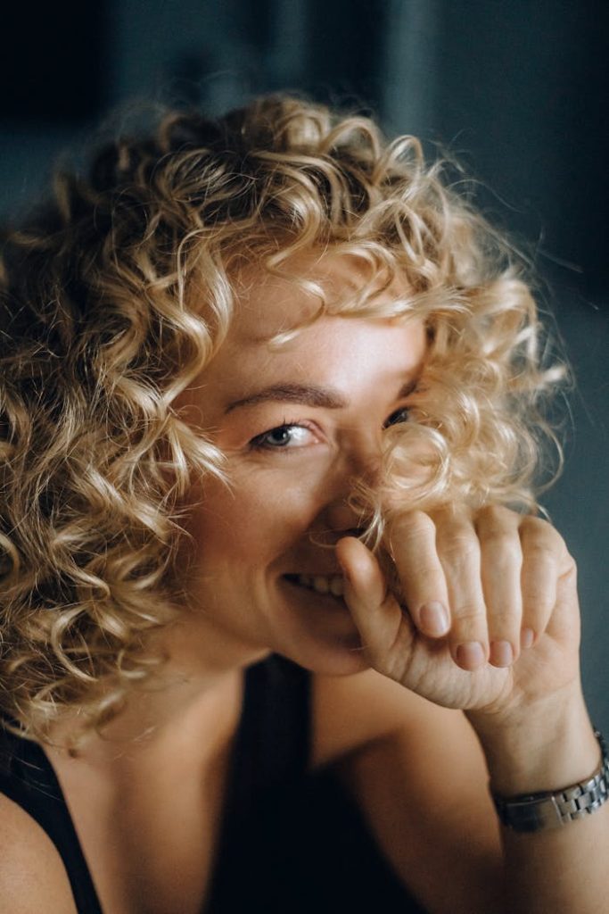 Close-up portrait of a cheerful woman with curly blonde hair covering her mouth playfully.
